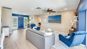 Una sala de estar moderna con un sofá gris, sillones azules, un ventilador de techo y un cuadro grande con una escena de playa.