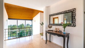 A spacious room with a wooden ceiling, a large window with a view of greenery, a console table with a mirror and decorative items, and a potted plant.