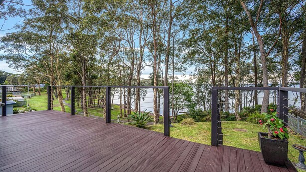 A wooden deck with a metal railing overlooking a lake, surrounded by trees and a grassy area. - WaterSide by Experience Jervis Bay (Sanctuary Point)