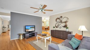 A modern living room with hardwood flooring, a ceiling fan, a flat-screen TV, a dining area with a table and chairs, and a decorative wall piece. - WaterSide by Experience Jervis Bay (Sanctuary Point)