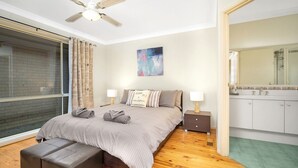 A bedroom with a bed, two bedside tables, a window with curtains, a ceiling fan, a painting, and a bathroom with a mirror and sink. - WaterSide by Experience Jervis Bay (Sanctuary Point)