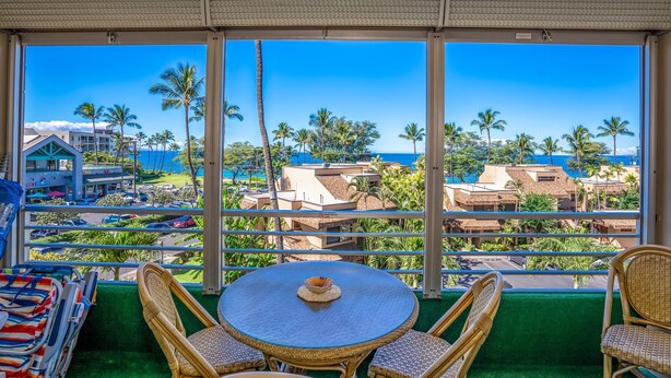 Een balkon met uitzicht op de oceaan en palmbomen, rieten stoelen, een ronde tafel met een kopje en een groen tapijt.