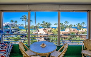 A balcony with a view of the ocean and palm trees, wicker chairs, a round table with a cup, and a green carpet.