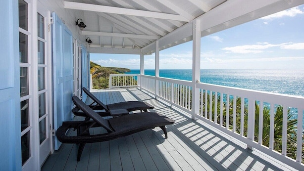Een overdekt terras met twee loungestoelen, een witte balustrade en uitzicht op zee.