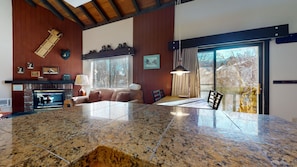Un salon spacieux avec une grande table en granit, une cheminée et une vue sur les arbres, visible à travers une porte-fenêtre coulissante.