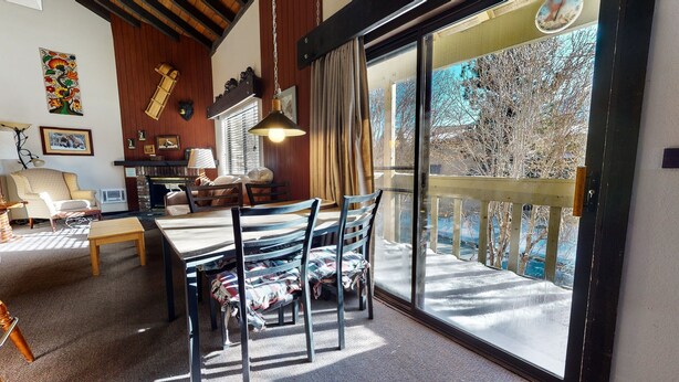 Una sala de estar acogedora con techo de madera, una mesa de comedor con sillas, una chimenea y un ventanal con vistas a árboles.