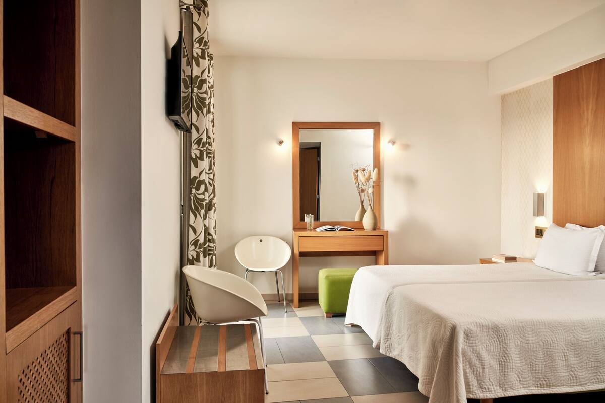 Quarto de hotel com uma cama, um guarda-roupa de madeira, uma penteadeira com espelho, uma cadeira e um banquinho.
