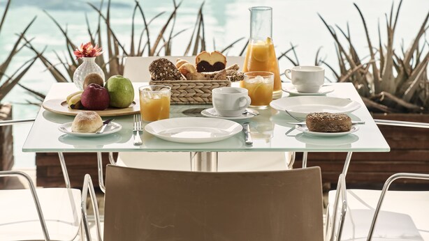 A breakfast table with various foods and drinks, set against a view of the ocean. - Oxygen Favie by GHH (Tinos)