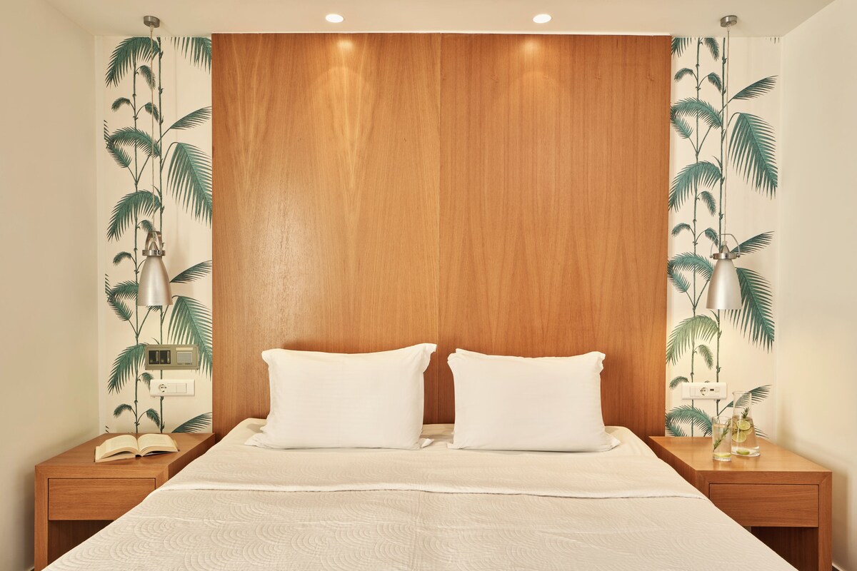 A bedroom with a wooden headboard, two white pillows, a bed with a textured white bedspread, a nightstand with a lamp, and a wall with a tropical leaf pattern.