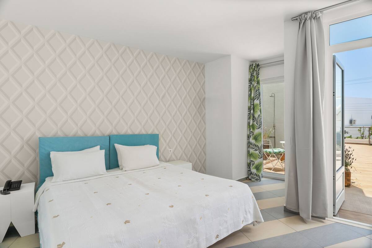 Quarto com cama, mesa de cabeceira, janela com cortinas e vista exterior.