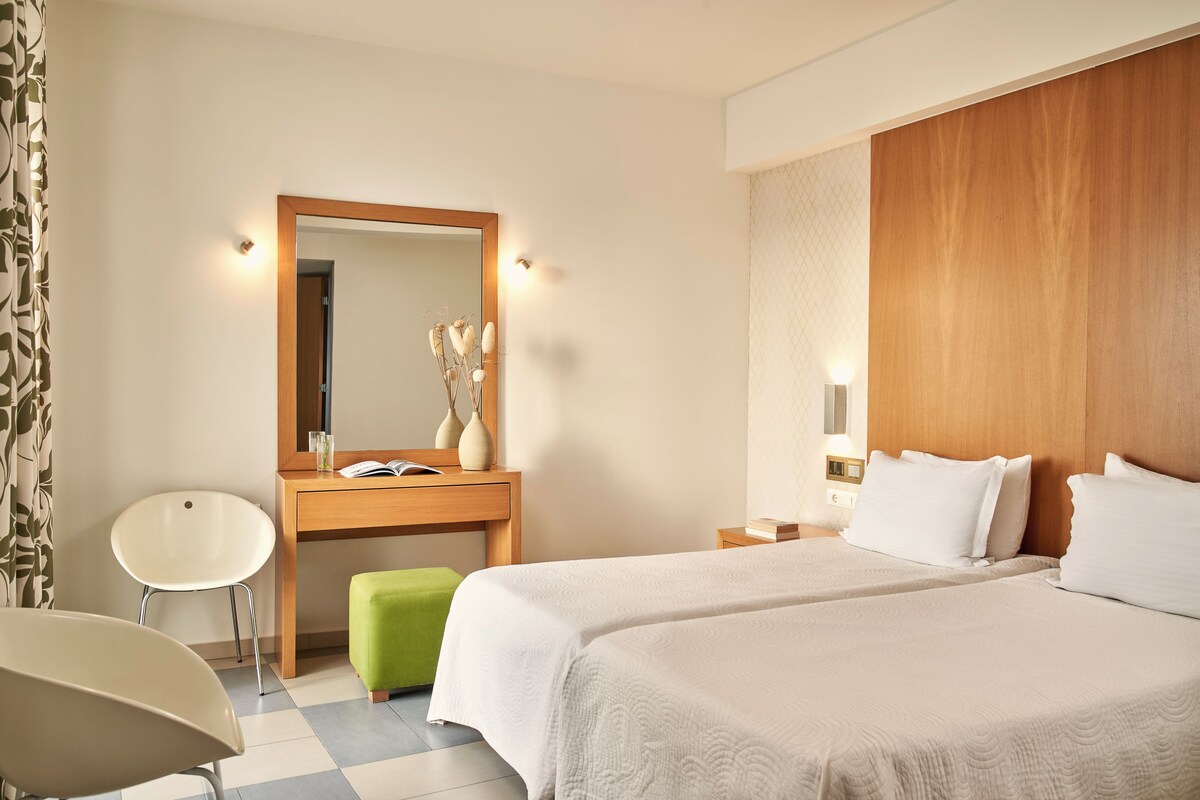 A hotel room with a wooden headboard, a large mirror, a desk with a chair, and a green ottoman.
