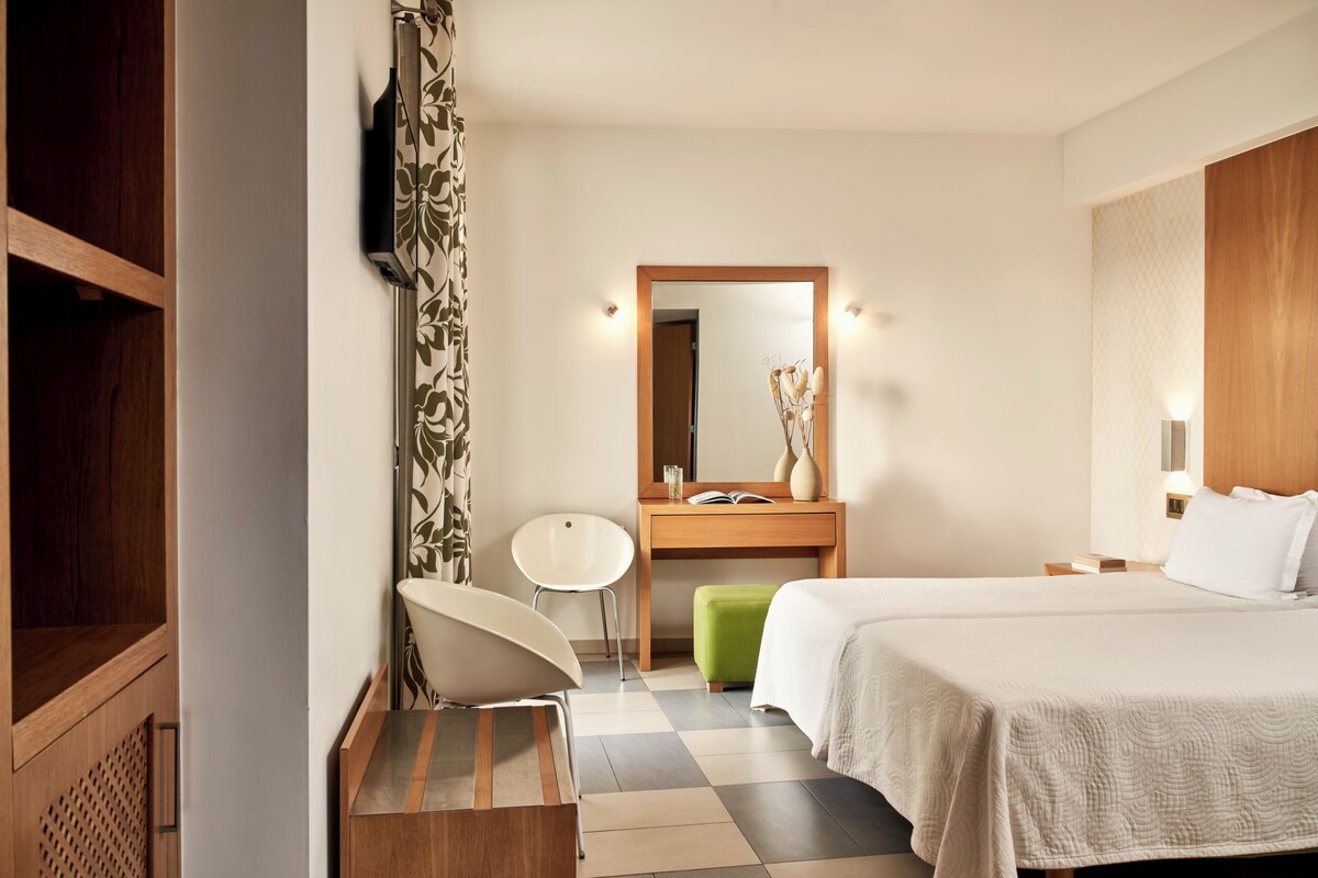 A hotel room with a bed, a wooden wardrobe, a vanity with a mirror, a chair, and a small table with a book and a vase.