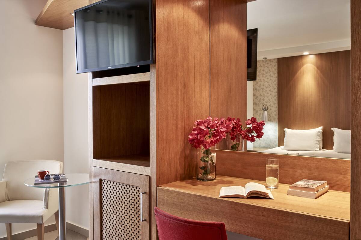 Quarto de hotel com escrivaninha de madeira, uma cadeira, televisão fixada na parede, cama com roupa de cama branca e um vaso com flores.