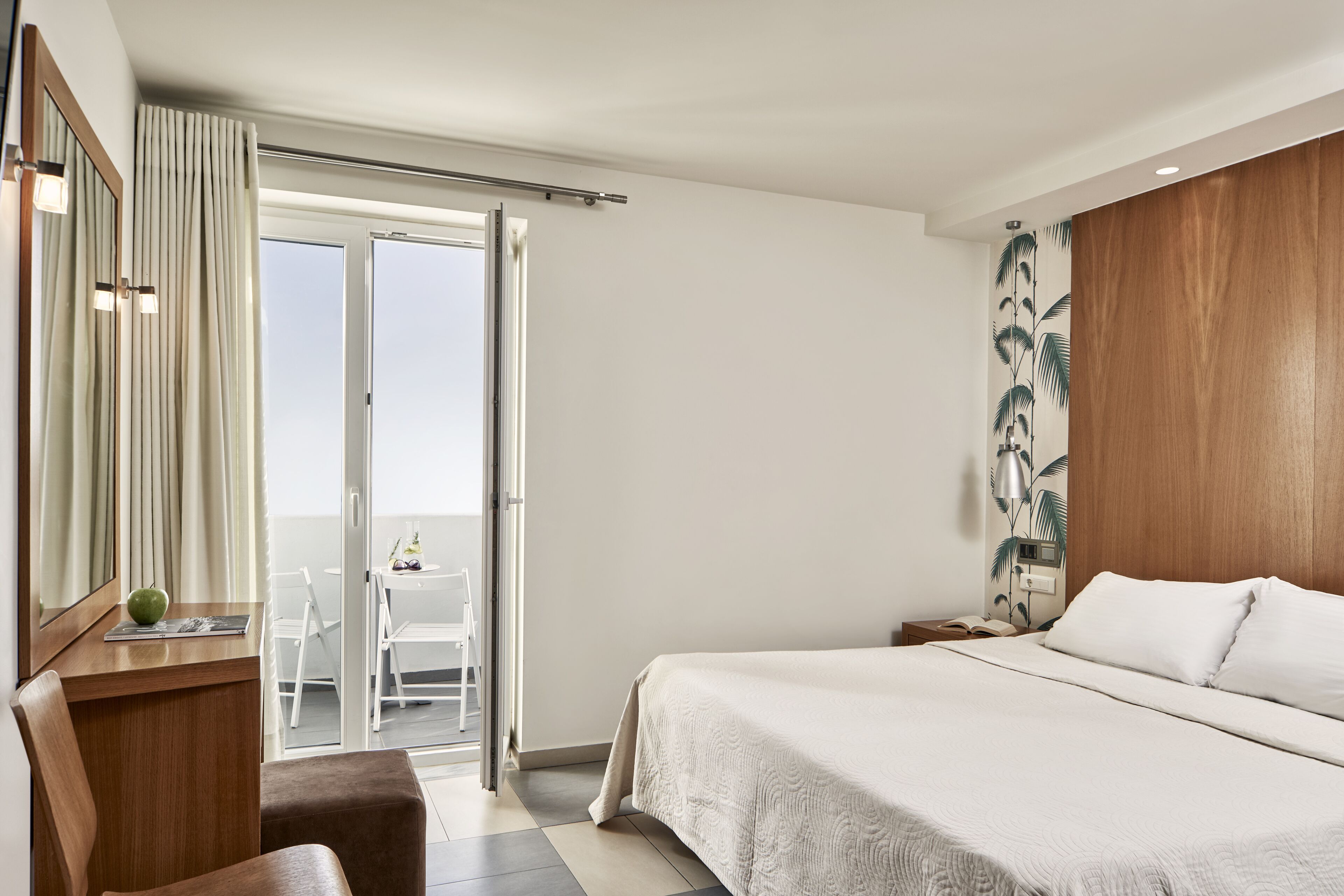 A hotel room with a bed, a desk, a chair, a mirror, a sliding glass door leading to a balcony, and a wall with a tropical plant mural.
