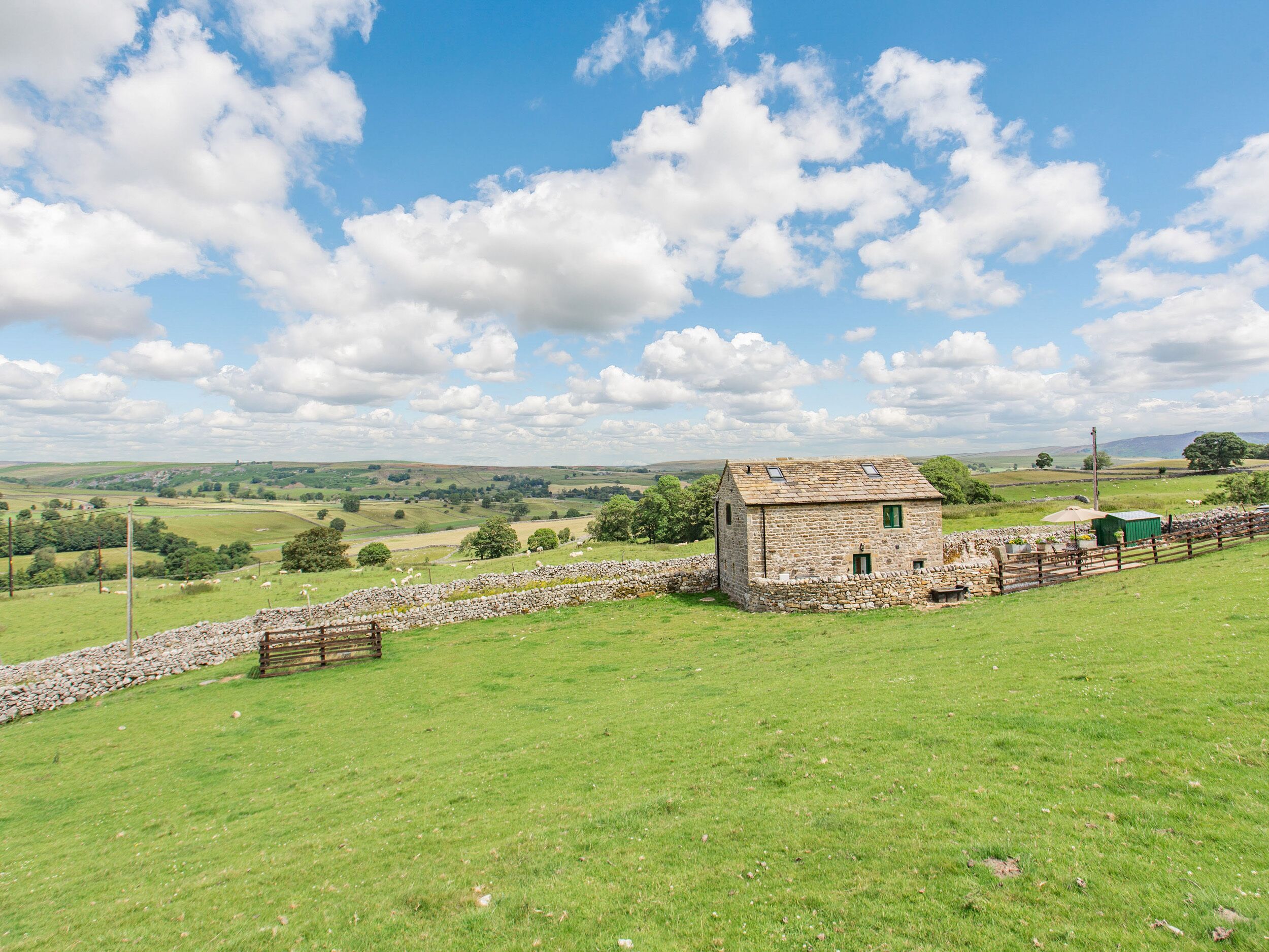 A stone cottage with a stone wall and a wooden fence, surrounded by a green field and a clear blue sky.