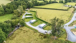 An aerial view of a rural property with several large, white, rectangular buildings, likely barns or storage units, arranged in a curved layout. There are fenced areas and a winding road connecting the buildings. - Oak Tree View (Llandrindod Wells)