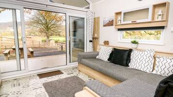 A modern living room with a grey sofa, patterned cushions, a wooden coffee table, and a large sliding glass door leading to an outdoor area with a wooden deck and a tree.