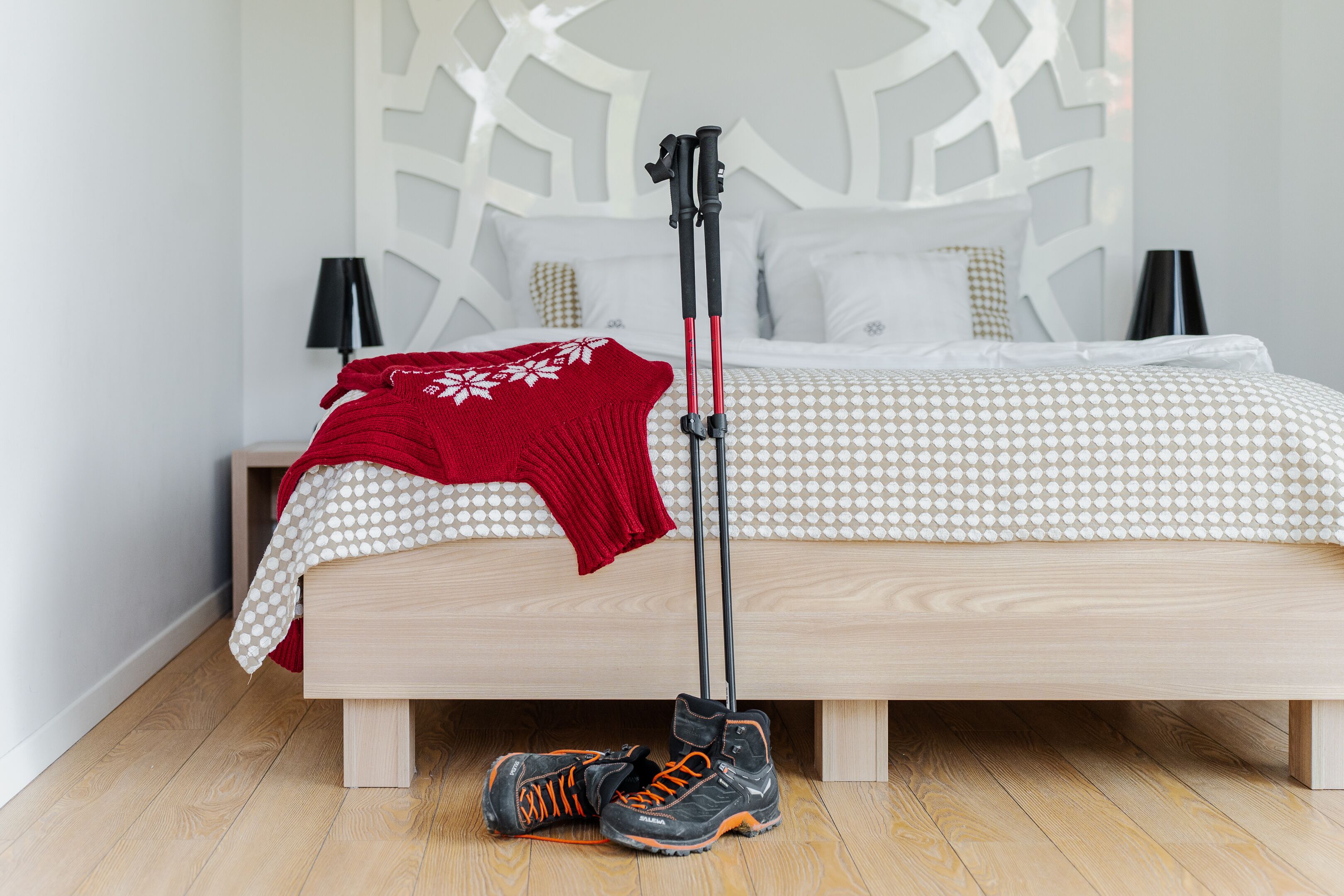 A neatly made bed with a patterned bedspread, a red throw blanket, and a pair of shoes and a tripod on the floor.