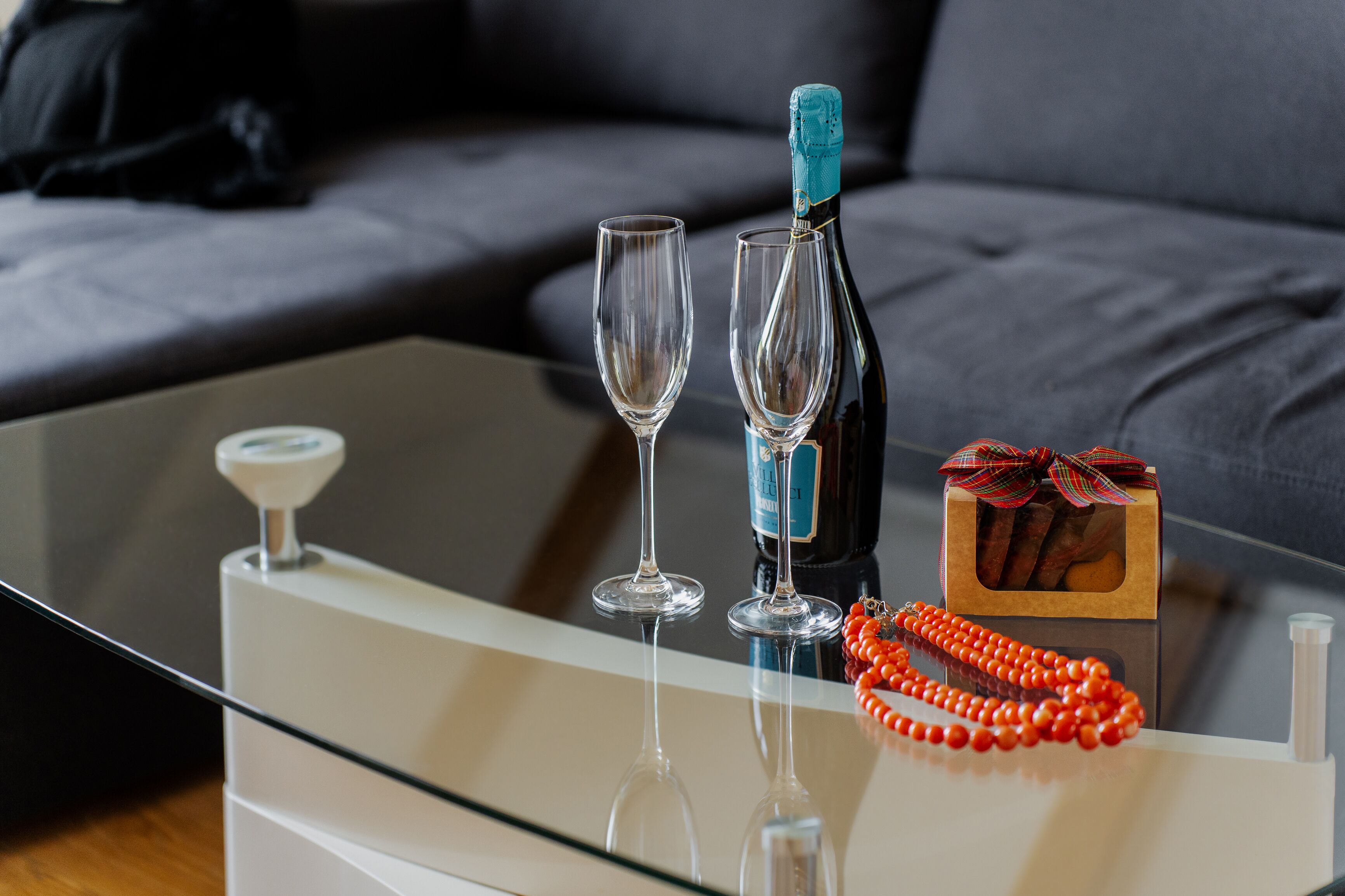Una mesa de vidrio con una botella de champán, dos copas de vino y una pequeña caja de regalo con un lazo.