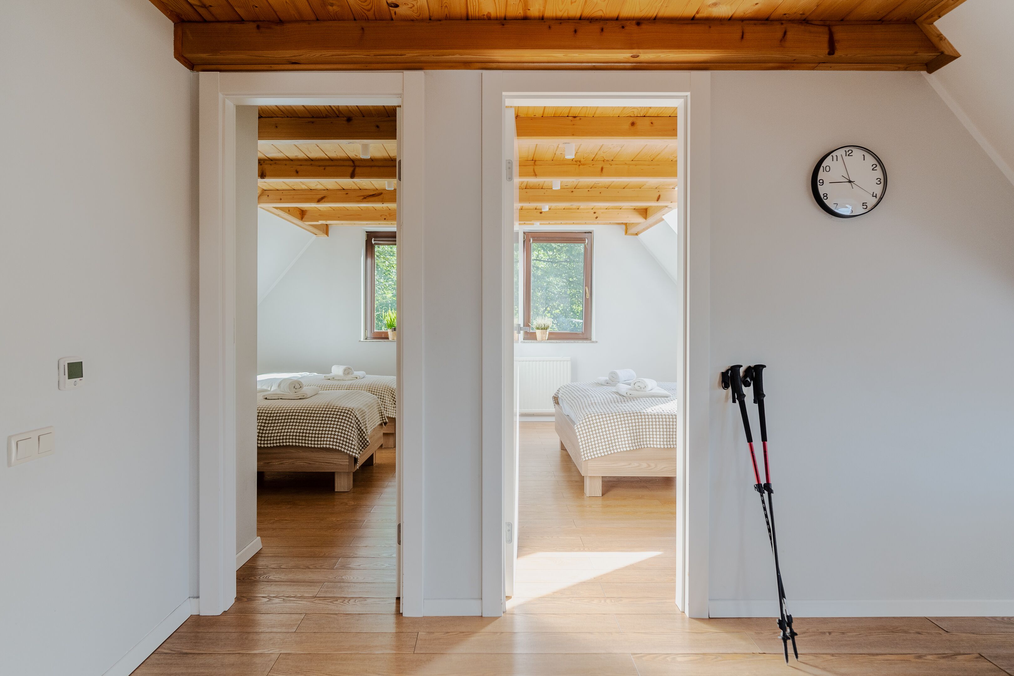 Una habitación con techo de madera, una cama con ropa de cama a cuadros, un reloj en la pared y un par de bastones de senderismo apoyados contra la pared.