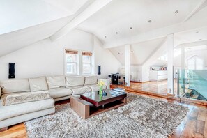 A spacious loft with a high ceiling, a large sofa, a coffee table, and a view of the kitchen through a glass door.
