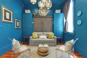 A room with a blue wall, a grey sofa, clear chairs, a wooden floor, and a large clock.