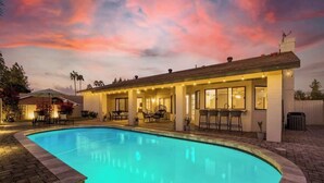 A swimming pool with a curved edge, a covered patio area with outdoor seating, and a building with large windows and a chimney. - Four BR Modern Tempe Oasis|Heated Pool + Game Room (Tempe)