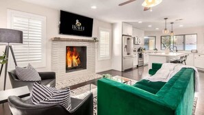 A modern living room with a fireplace, a ceiling fan, and a kitchen in the background. - Four BR Modern Tempe Oasis|Heated Pool + Game Room (Tempe)