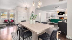 A spacious living room with a large wooden dining table, black leather chairs, and a green sofa. There is a fireplace and a pool table in the background. - Four BR Modern Tempe Oasis|Heated Pool + Game Room (Tempe)