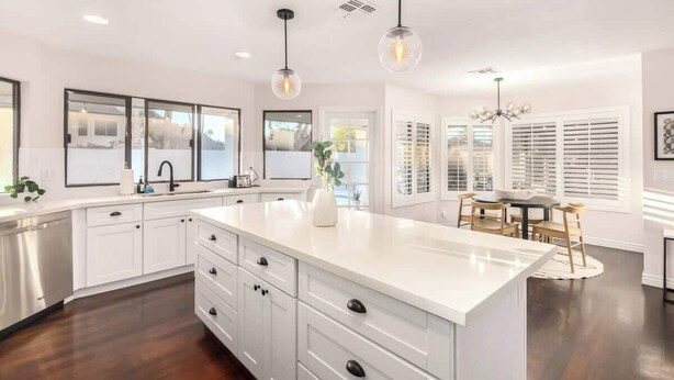 A modern kitchen with white cabinetry, a central island, pendant lighting, and a dining area with a table and chairs. - Four BR Modern Tempe Oasis|Heated Pool + Game Room (Tempe)