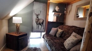 A cozy room with a wooden floor, a dark brown sofa with cushions, a small side table with a lamp, a wall-mounted shelf with decorative items, and a large painting of a deer. - Ground floor, parking, tv, ski locker, 61m², Valloire (Valloire)