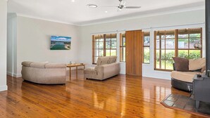 A spacious living room with wooden flooring, a ceiling fan, and large windows offering a view of greenery outside. - Minutes to Malua Bay Beach | Sunset Vista Retreat (Malua Bay)