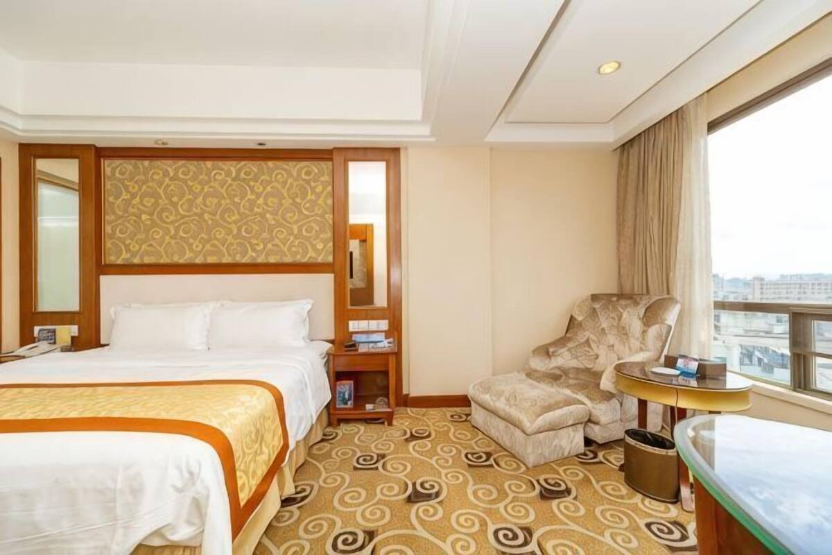 A hotel room with a bed, a chair, a small table, a nightstand, and a large window with a city view.