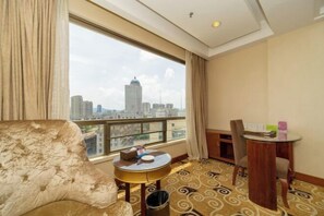 A hotel room with a large window offering a city view, a plush armchair, a small round table with a lamp, and a wooden chair. - Golden Diamond Hotel (Zhaoqing)