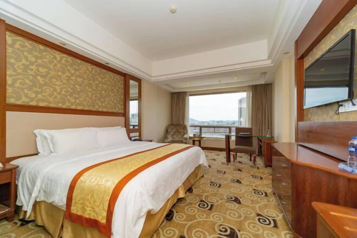 A hotel room with a large bed, a desk, a chair, a TV, and a view of the city.