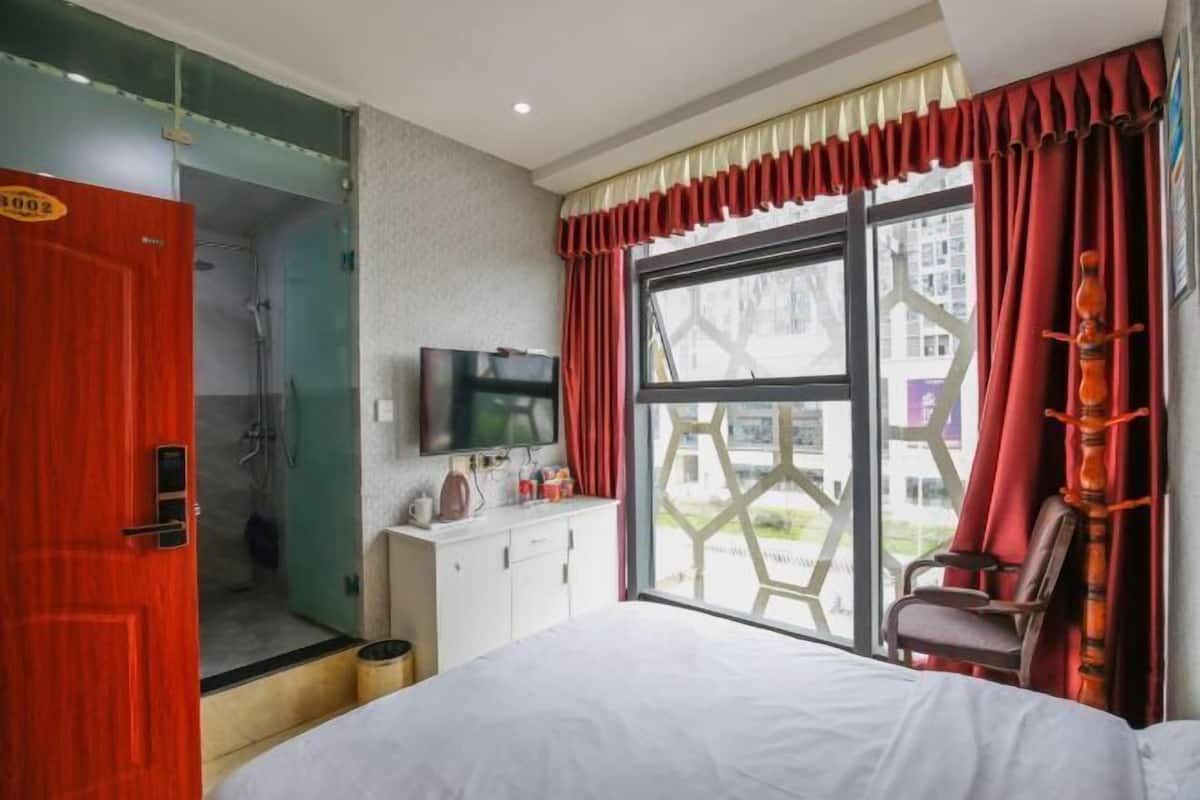 Una habitación de hotel con cama, televisión, escritorio con silla, ventana con cortinas y zona de ducha.