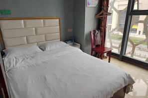 Habitació d'hotel amb un llit gran, un panell de capçalera de fusta, una nitòria, una cadira vermella i una finestra amb vista a edificis.