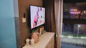 A room with a television mounted on the wall, a small table with a kettle and cups, and a window with a view of a cityscape at night. - Guang'an Yandu Hotel (Guang'an)