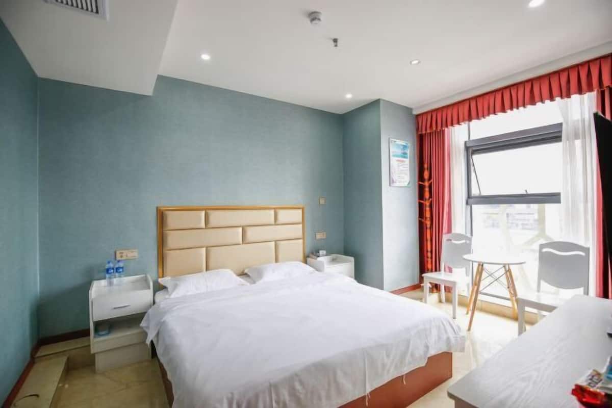 Una habitación de hotel con cama, mesitas de noche, una silla, una pequeña mesa y un ventanal con cortinas.