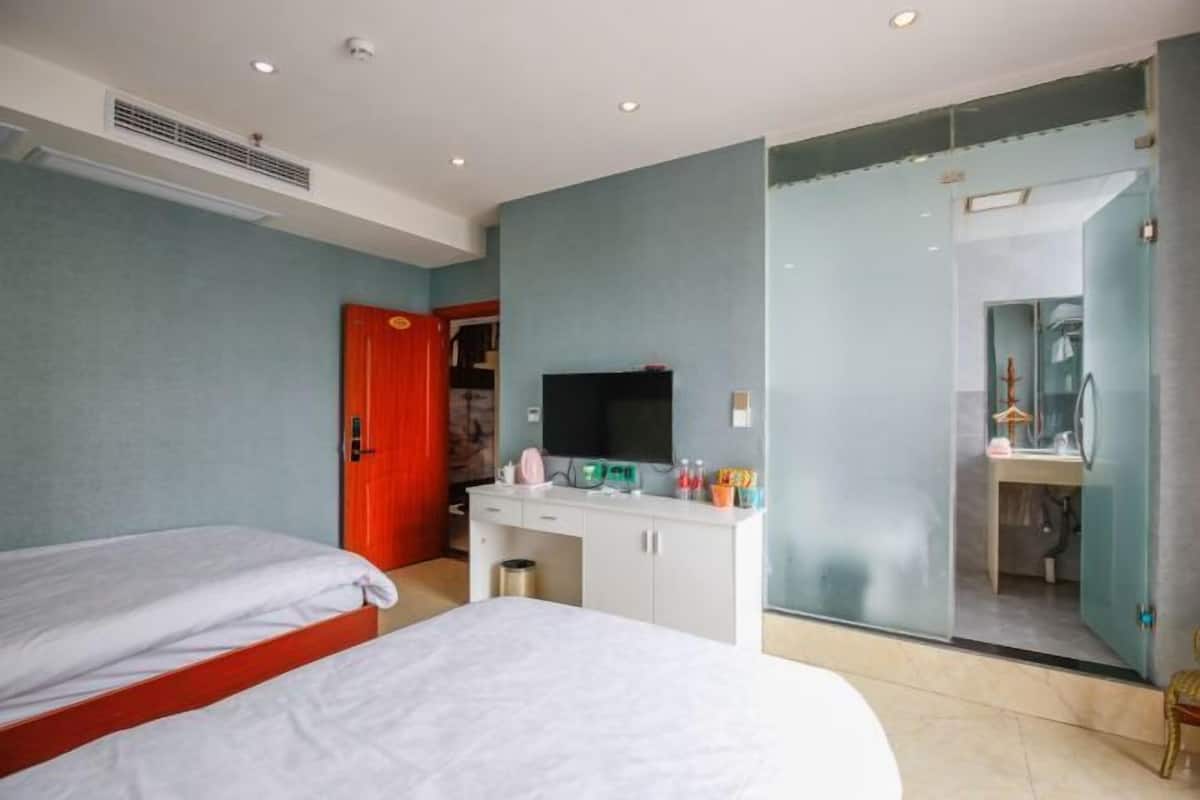 Habitación de hotel con dos camas, un escritorio con televisor y un baño visible a través de una puerta de vidrio.