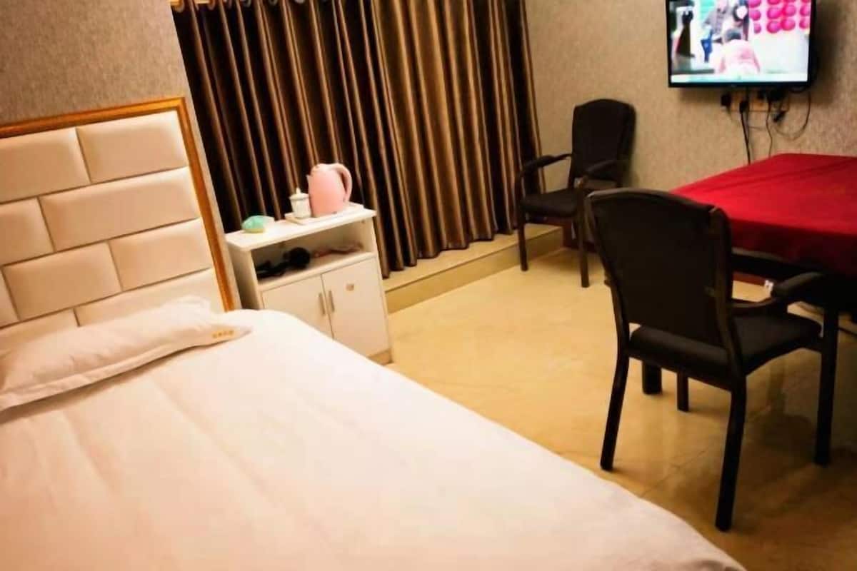 Una habitación de hotel con una cama, una mesita de noche con una tetera rosa, una silla, un televisor montado en la pared y una mesa cubierta con mantel rojo.