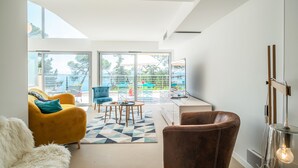 Ett modernt vardagsrum med en gul fåtölj, en brun fåtölj och en vit, lurvig stol. Ett glasbord står framför en geometrisk matta. På höger sida finns ett glasskåp och en lampa med en synlig glödlampa. Stora fönster ger utsikt över omgivningen.