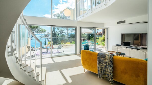 En modern inredning med en gul soffa, en tv och en trappa som leder upp till en balkong med utsikt över havet.