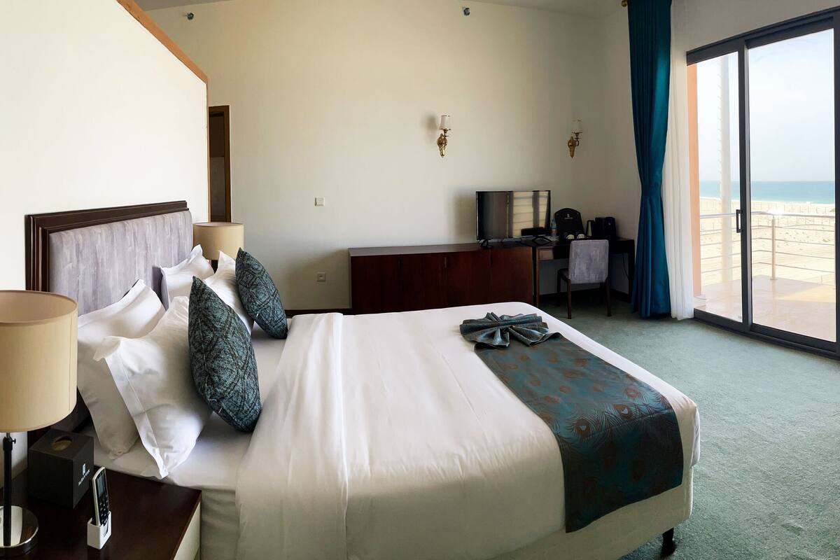 A hotel room with a bed, a desk, a chair, a lamp, and a view of the beach.