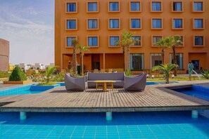 A pool area with a wooden deck, lounge chairs, and a building with multiple windows in the background. - Hotel Al Salam Resort (Nouakchott)