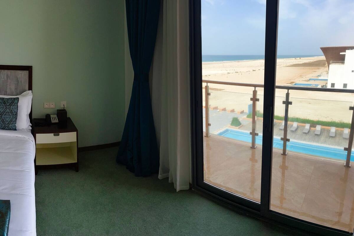 A hotel room with a bed, a TV, a desk, and a view of the beach.