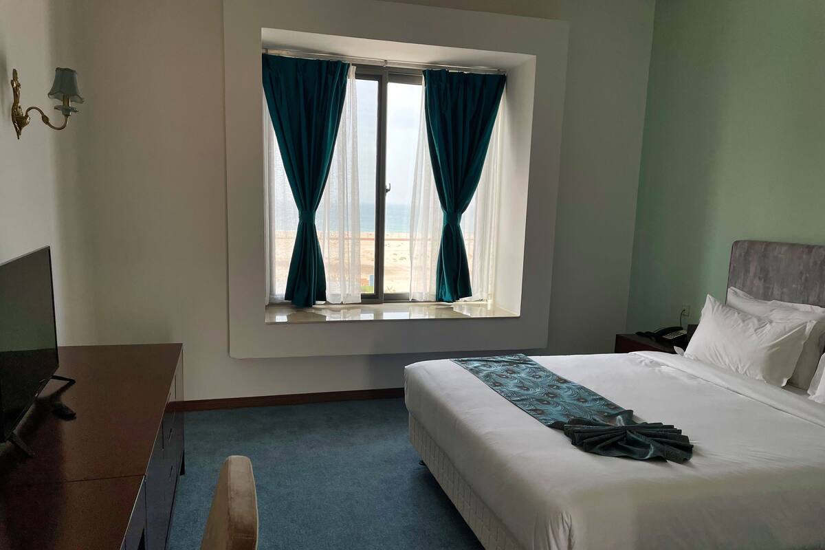 A hotel room with a bed, a desk, a chair, a window with curtains, and a view of the beach.