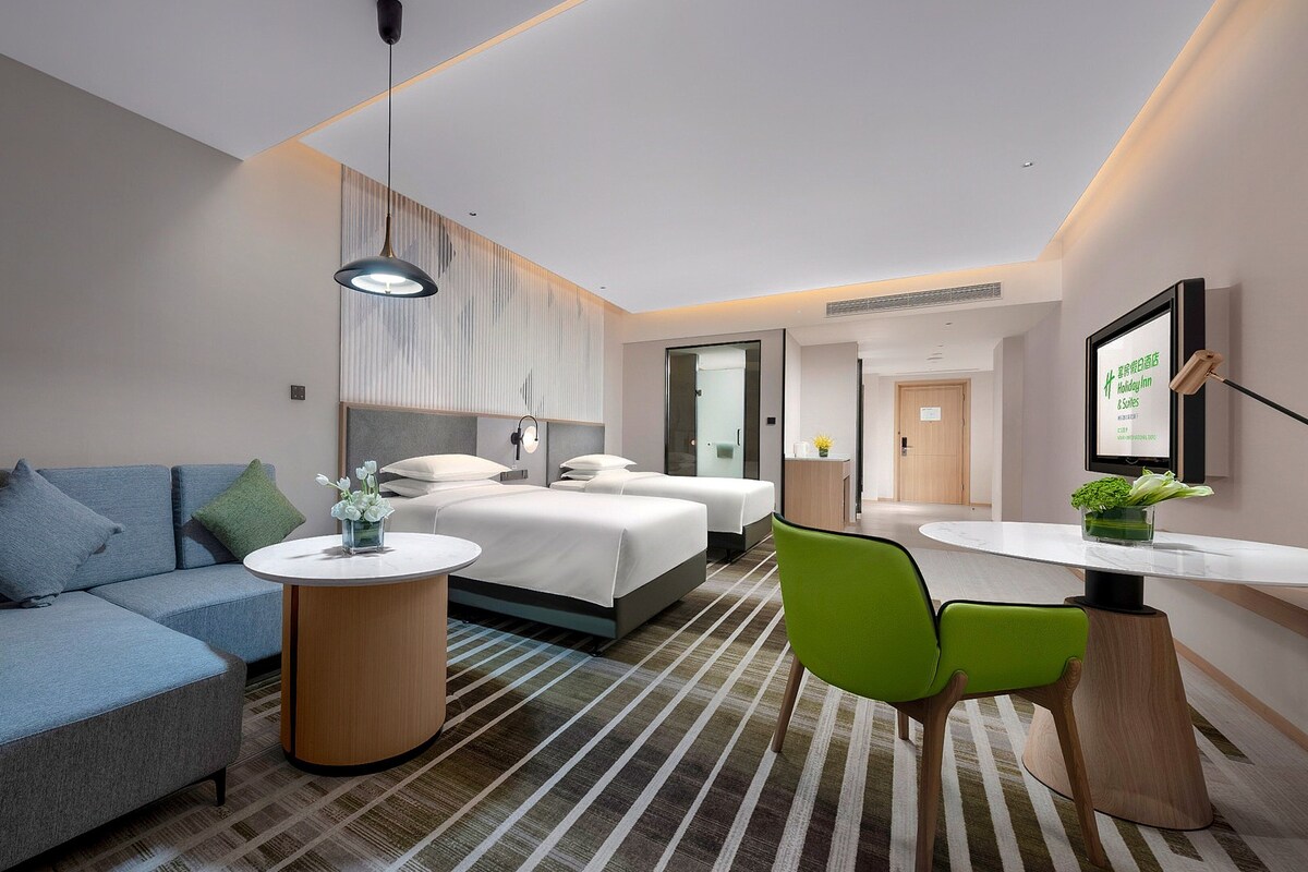 A modern hotel room with a sofa, two beds, a dining table, and a green chair.