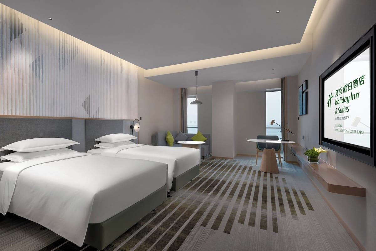 Una habitación de hotel moderna con dos camas, un escritorio y una pantalla grande mostrando información del hotel.