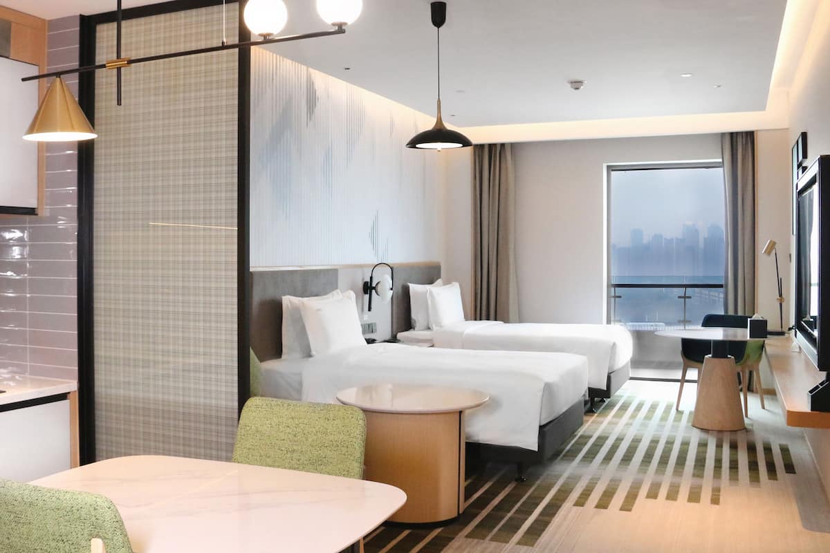 Una habitación de hotel moderna con kitchenette, dos camas, zona de comedor y un ventanal con vistas a la ciudad.